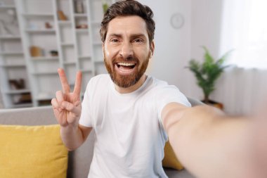 Selfie çeker, video görüşmesi yapar, barış sembolü gösterir, neşeli genç adam sıcak oturma odasında selfie çeker. Sarı yastıklarla çevrili, parlak bir şekilde gülümserken bir eliyle barış işareti yapıyor.
