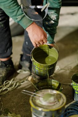 Bir insan, loş bir atölyede yaratıcı bir girişime hazırlanırken canlı yeşil boya döken bir boya kutusu tutuyor. Etrafa çeşitli boya kutuları saçılmış..