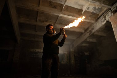 Koruyucu giysili bir birey, sahipsiz bir yapının içinde bir alev makinesini tutuşturur ve karanlık alana turuncu bir parıltı saçar. Çevre, heyecan ve keşif hissi uyandırıyor..