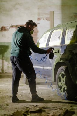 Maske spreyi kullanan biri eski bir atölyenin içine araba boyuyor. Çevre tozlu ve endüstriyel, araba özelleştirmesindeki odak ve çabayı vurguluyor.