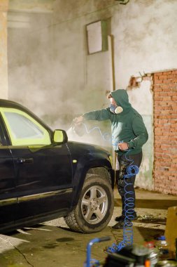 Kapüşonlu ve güvenlik maskeli biri loş bir endüstriyel garajda siyah bir araca boya sıkar. Atmosfer sprey boyadan gelen sisle dolu..