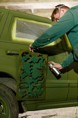 Bir kişi bir araç üzerinde çalışıyor, sprey boyayla kamuflaj desenleri eklemek için stencil kullanıyor. Dekor iyi aydınlatılmış bir garaj, araba için benzersiz bir özelleştirme projesi sunuyor..