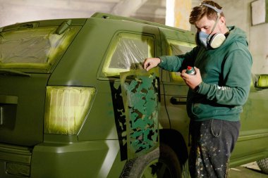 Bir kişi maske ve koruyucu giysiler giyerken yeşil bir SUV 'ye kamuflaj desenleri uyguluyor. Dekor, yakınlarda boya malzemeleri olan iyi aydınlatılmış bir depo..