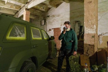 Bir işçi, sıcak ışıkla aydınlatılmış loş, endüstriyel bir alanda kamuflaj renklerine sahip büyük bir aracı dikkatlice boyarken maske takıyor. Karton parçalar ve aletler yakında..