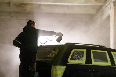 Bir insan gece loş bir endüstriyel atölyede sprey boyayla araba boyuyor. Puslu hava ve koruyucu kaplamalar, araç yeni bir palto için hazırlanırken sanatsal bir atmosfer yaratıyor..