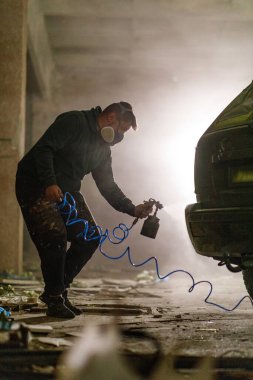 Terk edilmiş bir depoda maske takan bir adam bir araca boya sıkar. Loş ışık ve toz, projesinde titizlikle çalışırken dengesiz bir atmosfer yaratıyor..