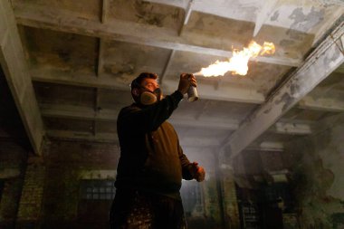 Gaz maskesi takan bir kişi sprey kutusunu ateşler, ıssız bir depoda dramatik bir alev üretir, loş bir çevrede yaratıcılık ve tehlikenin bir karışımını sergiler..
