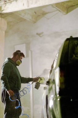 Koruyucu maske takan bir kişi, hafif aydınlatılmış bir garajda el bilgisayarı ile bir arabaya dikkatlice püskürtüyor ve pürüzsüz bir kapanış yapmaya odaklanıyor. Atmosfer sprey sisi ile dolu..