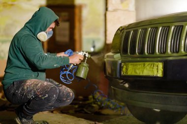 Bir insan loş bir atölyede sprey tabanca kullanarak araç boyamaya odaklanır. Kapüşon ve maske takıyorlar, açıkça ön taraftaki arabaların tamiri üzerinde çalışıyorlar..