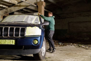 Bir işçi terkedilmiş bir depoda tadilat için bir Jeep hazırlıyor. Araç maskeleme bandıyla kaplı ve enkaz loş alana saçılmış durumda..