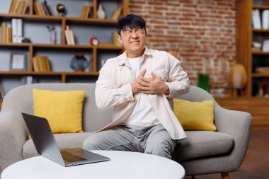 Çalışan, kalp krizi geçiren, göğsü ağrıyan bir adam, gri bir koltuğa oturmuş, göğsünü rahatsız eden parlak sarı yastıklarla çevrili. Sıcak ve davetkar bir oturma odasında dizüstü bilgisayara bakarken endişeli görünüyor..