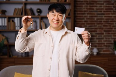 Satın alma odası kartı, daire anahtarları, kredi kartı, gülümseyen adam, neşeli adam bir elinde bir şişe, diğer elinde kimlik kartı, kitaplarla dolu karşılama odasında duruyor ve rahat bir kanepe.