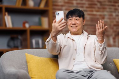 Video konuşması, telefon konuşması, gülümseyen Asyalı adam modern ev dekorasyonuyla çevrili rahat bir kanepeden gelen görüntülü bir aramayla gülümsüyor. Güneşli bir öğleden sonra, davetkar bir atmosfer yaratıyor..