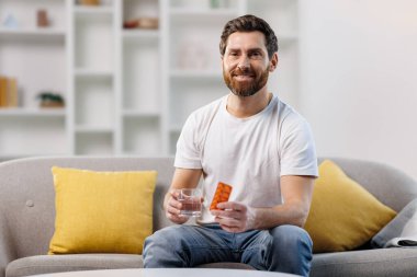 Hap alan adam, hasta adam, baş ağrısı çeken, sakallı genç adam, sarı yastıklı bir kanepede rahatça oturuyor, bir elinde takviye paketi, diğer elinde bir bardak su..