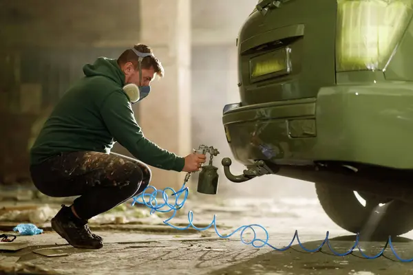 Bir zanaatkar, korunmak için maske takarak ıssız bir depoda aracın arkasına boya sürer. Çalışma alanı enkaz ve gölgelerle dolu, odaklandığı işi vurguluyor..