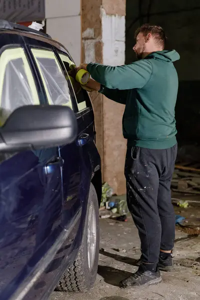 Loş bir oto tamirhanesinde özenle çalışan bir adam, resme hazırlanmak için maske bandını dikkatlice mavi bir arabaya takar ve görevinde hassasiyet sağlar..
