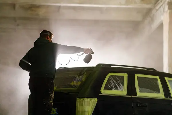 Bir insan gece loş bir endüstriyel atölyede sprey boyayla araba boyuyor. Puslu hava ve koruyucu kaplamalar, araç yeni bir palto için hazırlanırken sanatsal bir atmosfer yaratıyor..