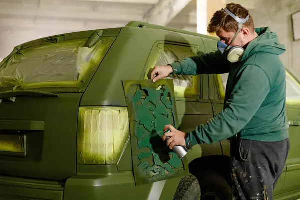 Bir kişi atölyede kamuflaj desenli bir SUV 'yi spreyle boyuyor. Maske takarak, özgün ve koruyucu bir tasarım yaratmak için şablon kullanarak dikkatlice boya sürüyorlar..