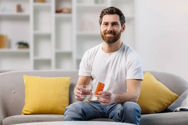 Hap alan adam, hasta adam, baş ağrısı çeken, sakallı genç adam, sarı yastıklı bir kanepede rahatça oturuyor, bir elinde takviye paketi, diğer elinde bir bardak su..