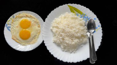 Bir tabak dolusu pişmiş beyaz pirinç çifte güneşli yan yumurtalarla birlikte siyah arka planda çatal ve kaşık görüntüsü. 