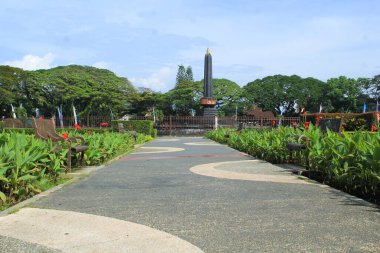 Balai kota malang parkının kaldırımı kenarında yeşil çalılık ve Tugu malang anıtı arka planında
