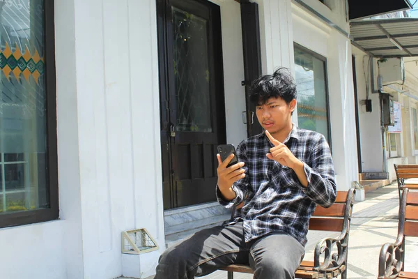 当有人用智能手机打电话 坐在木制长椅上 愤怒的亚洲年轻人指指点点 进行威胁 — 图库照片