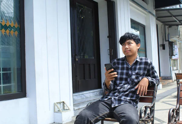 一个穿着蓝色法兰绒衬衫的不快乐的亚洲年轻人拿着黑色智能手机 神智不清 神智不清 — 图库照片