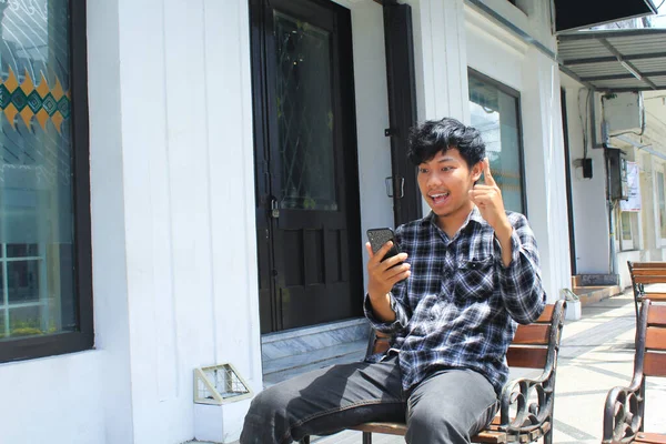 当看到自己的智能手机 坐在白色建筑窗户旁边的木制长椅上的时候 这位兴奋的亚洲年轻人有了一个绝妙的主意 — 图库照片