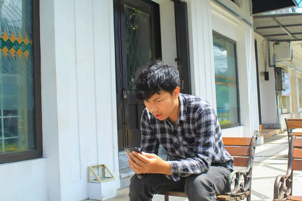 年轻的亚洲男子穿着蓝色法兰绒衬衫 坐在白色建筑旁边的木制长椅上 震惊而惊讶 — 图库照片