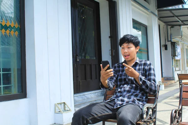 身穿蓝色法兰绒衬衫 坐在白色建筑旁边长椅上的兴奋的亚洲年轻人微笑着 指着他的智能手机 — 图库照片