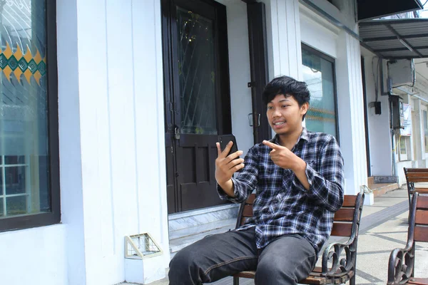身穿蓝色法兰绒衬衫 坐在白色建筑旁边长椅上的兴奋的亚洲年轻人微笑着 指着他的智能手机 — 图库照片