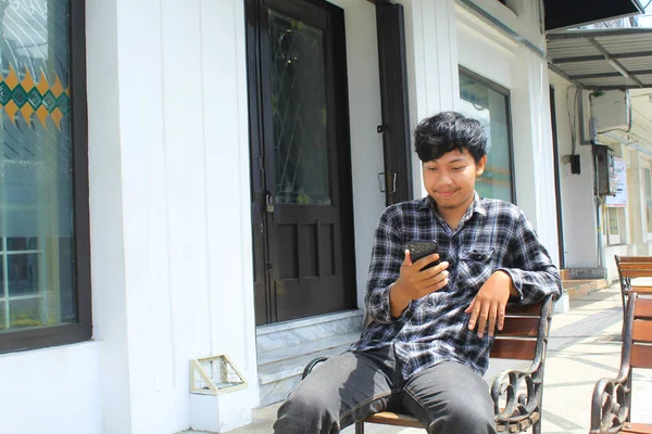 一个快乐的亚洲年轻人看到他的黑色智能手机时 面带微笑 一个快乐的青少年穿着法兰绒衬衫坐在白色和黑色建筑旁边的长椅上 面带微笑 — 图库照片