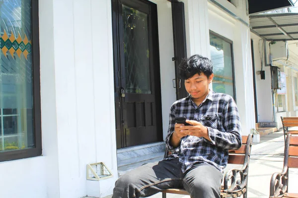当一个快乐的亚洲年轻人用他的黑色智能手机发短信时 他笑了 一个快乐的青少年穿着法兰绒衬衫坐在白色和黑色建筑旁边的长椅上 面带微笑 — 图库照片