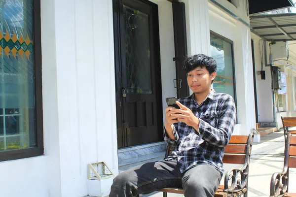当一个快乐的亚洲年轻人用他的黑色智能手机发短信时 他笑了 一个快乐的青少年穿着法兰绒衬衫坐在白色和黑色建筑旁边的长椅上 面带微笑 — 图库照片