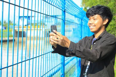 Asyalı genç adam gülümsüyor ve tatilde selfie çekiyor. Siyah ceketli genç adam demir çitin yanında duruyor ve heyecanlı bir yüzle video arıyor.