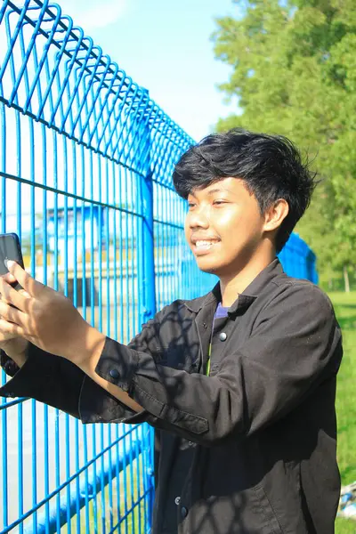 アジア系の青年は休暇中に自撮り写真を撮っています 黒いジャケットを着た若者が鉄の柵の横に立って興奮した顔でビデオ通話をしています — ストック写真
