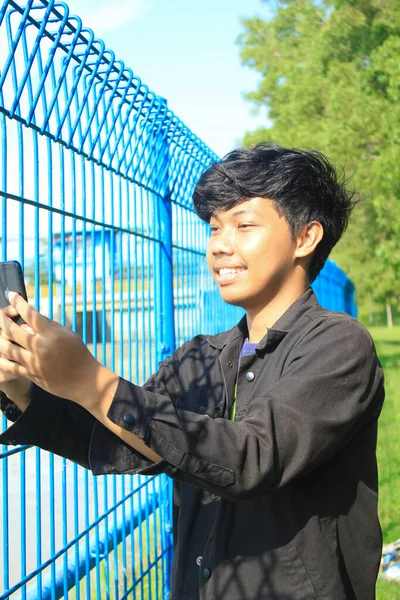アジア系の青年は休暇中に自撮り写真を撮っています 黒いジャケットを着た若者が鉄の柵の横に立って興奮した顔でビデオ通話をしています — ストック写真
