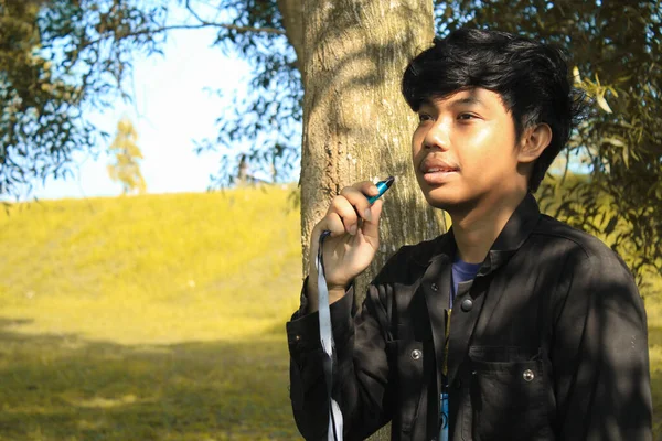 坦率而快乐的年轻亚洲男子拿着蓝色的蒸气装置 用黑白相间的带着树木和草地背景的灯笼来代替吸烟 穿着黑色夹克的放松的年轻人在笑 — 图库照片