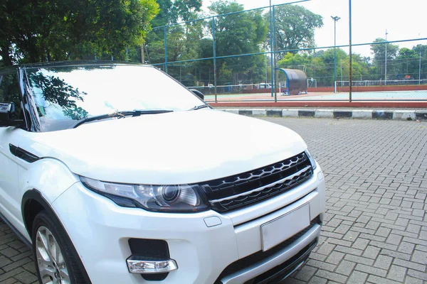 stock image front view of white crossover car parked beside of tennis court. suv car with and green trees