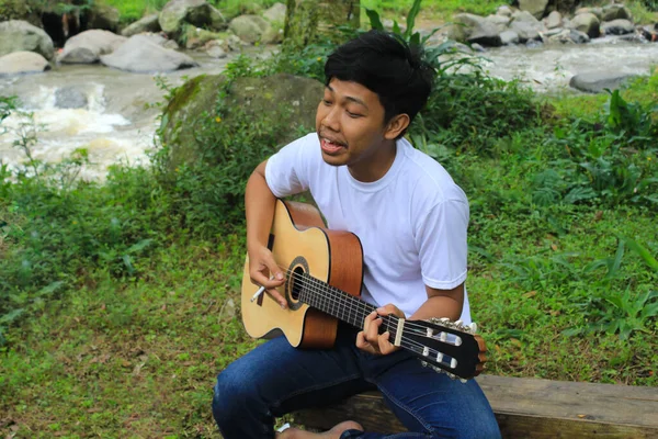 当假日野餐时 快乐的亚洲年轻人正在河边弹吉他 年轻的印度尼西亚男子旅行者在营地里弹奏吉他 — 图库照片