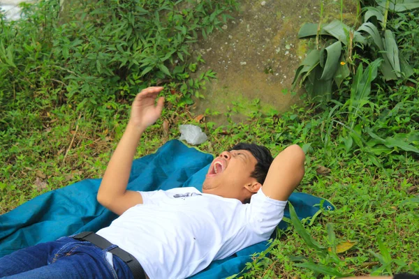 young man asian is lying on the lawn and yawn. young man relaxing by lying on the grass ground and yawning wearing white t shirt