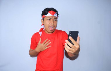 Endonezyalı genç adam Endonezya 'nın bağımsızlık gününde kırmızı tişört ve saç bandı takarak cep telefonundan sürpriz haberler izledikten sonra şok oldu.