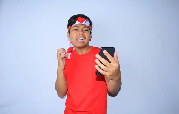 フリーランスのインドネシアの男は 彼の暴徒の携帯電話からメッセージを受け取った後怒っている赤いTシャツとヘッドバンドを身に着けています 孤立した白 — ストック写真