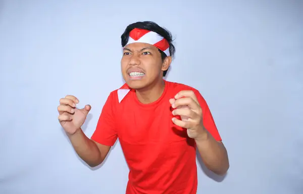 怒っているインドネシアの男は激しく叫んで インドネシア独立記念日に赤いTシャツとヘッドバンドを着て手を絞っている — ストック写真