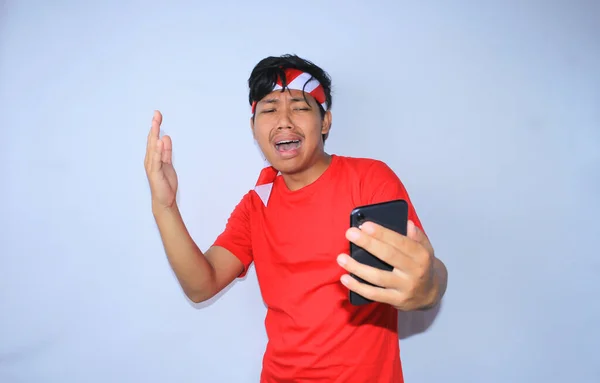 悲しいインドネシアの若者は スマートフォンを見ると泣いていて 白で隔離された赤いTシャツとヘッドバンドを着ています — ストック写真