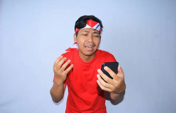 悲しいインドネシアの若者は スマートフォンを見ると泣いていて 白で隔離された赤いTシャツとヘッドバンドを着ています — ストック写真