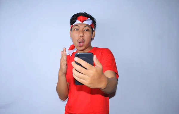 幸せなインドネシアの若者はニュースに驚いた彼の携帯電話を見てショックを受け インドネシア独立記念日に赤いTシャツとヘッドバンドを着て — ストック写真