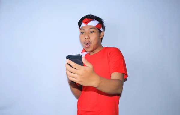 幸せなインドネシアの若者はニュースに驚いた彼の携帯電話を見てショックを受け インドネシア独立記念日に赤いTシャツとヘッドバンドを着て — ストック写真