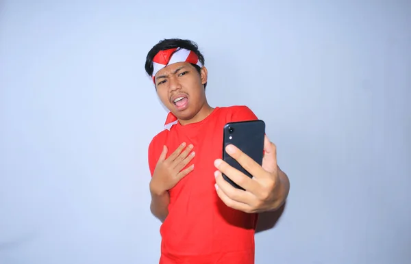 インドネシアの若い男性は 彼の携帯電話で驚いたニュースを見てショックを受け インドネシア独立記念日に赤いTシャツとヘッドバンドを着ていました — ストック写真