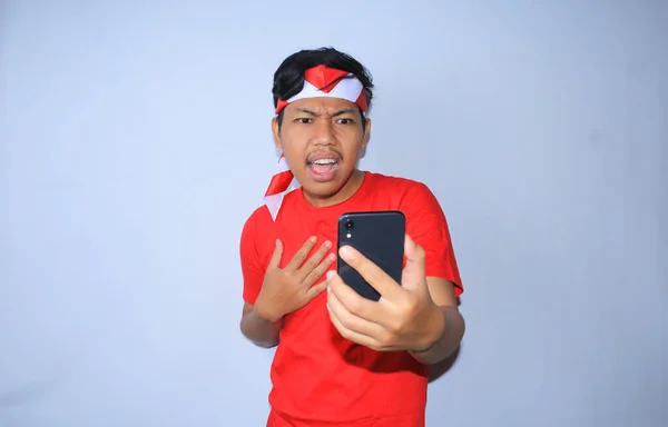 インドネシアの若い男性は 彼の携帯電話で驚いたニュースを見てショックを受け インドネシア独立記念日に赤いTシャツとヘッドバンドを着ていました — ストック写真
